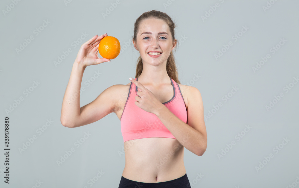 Beautiful young woman pointing finger at an orange fruit in her hand while standing against a gray background and looking directly at the camera. Beautiful young woman pointing finger at an orange fruit in her hand while standing against a gray background and looking directly at the camera.