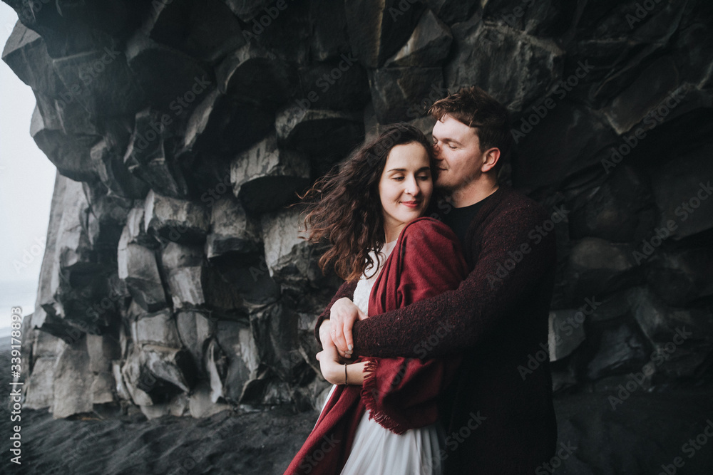 Wedding in Iceland. A young guy and a girl in a white dress and a red sweater are hugging near the black rock