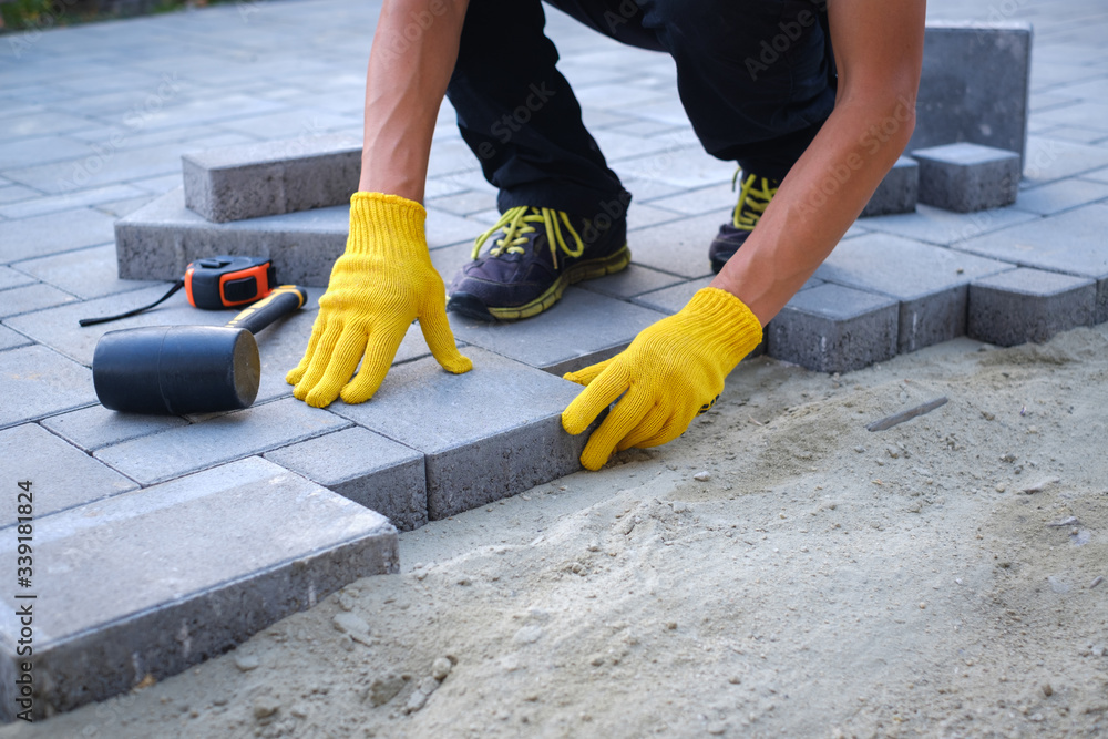 The master in yellow gloves lays paving stones in layers. Garden brick pathway paving by professional paver worker. Laying gray concrete paving slabs in house courtyard on sand foundation base. The master in yellow gloves lays paving stones in layers. Garden brick pathway paving by professional paver worker. Laying gray concrete paving slabs in house courtyard on sand foundation base.