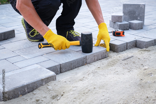 The master in yellow gloves lays paving stones in layers. Garden brick pathway paving by professional paver worker. Laying gray concrete paving slabs in house courtyard on sand foundation base.