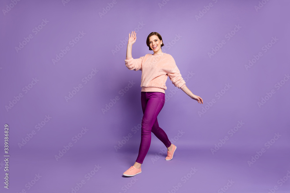 Full body photo of pretty lady walking down street good mood nice sunny day waving palm saying hi wear casual stylish pink jumper pants isolated purple color background Full body photo of pretty lady walking down street good mood nice sunny day waving palm saying hi wear casual stylish pink jumper pants isolated purple color background