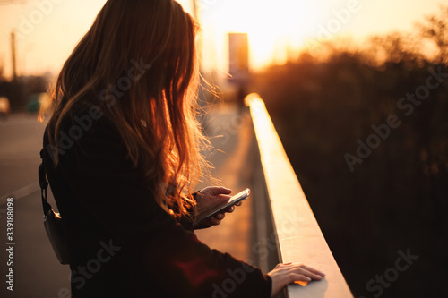 Young woman using smart phone while holding railing standing on bridge in city at sunset