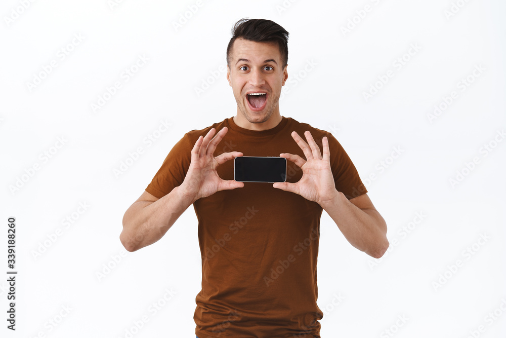 Excited smiling caucasian handsome man, holding smartphone over chest and showing cell screen, introduce new application on mobile phone, product update, new shopping online site, white background Excited smiling caucasian handsome man, holding smartphone over chest and showing cell screen, introduce new application on mobile phone, product update, new shopping online site, white background