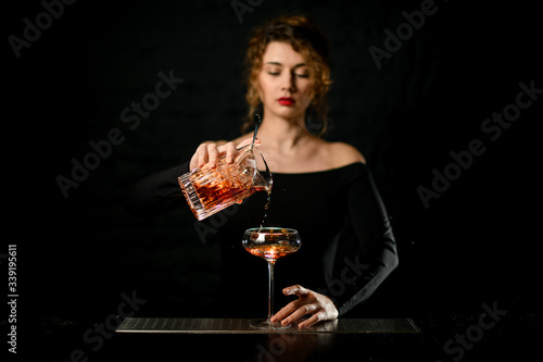 Woman bartender in dark bar carefully pours cocktail into wineglass.