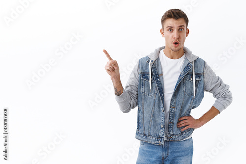 Portrait of curious handsome caucasian blond guy asking question about interesting advertisement offer, pointing finger upper left corner, open mouth excited, look camera white background