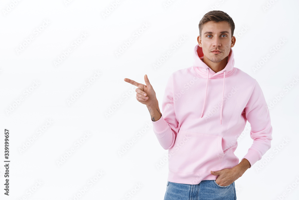 Portrait of handsome blond man in pink hoodie, pointing finger left at blank space for your promo, recommend visit online shop, click link, showing way to event, smiling camera, white background Portrait of handsome blond man in pink hoodie, pointing finger left at blank space for your promo, recommend visit online shop, click link, showing way to event, smiling camera, white background