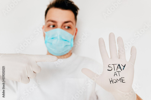 A young european man in blue mask shows a message Stay at home on gloves. Corona virus protection. Quarantine for Covid-19 prevention