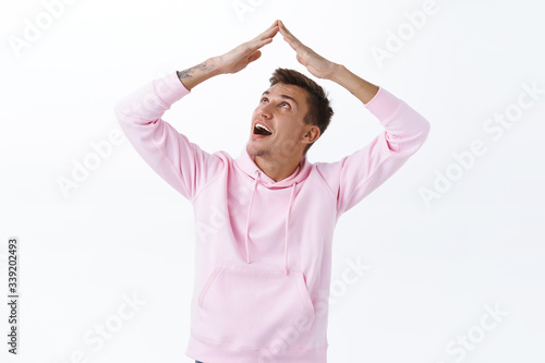 Stay home, self quarantine concept. Portrait of funny handsome blond boyfriend in pink hoodie, maiking roof with hands over head, isolating at house during covid19 epidemic, white background photo