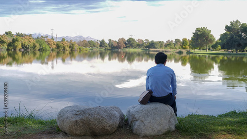 A pensive man sitting on a rock by the lake in the park, back view