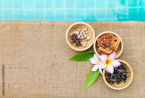 Thai dry herbs in round wooden bowl with Plumeria flower on hessian fabric background, spa concept, Dry Bael fruit and tea with Thai spice in wooden cup with space on blue water background