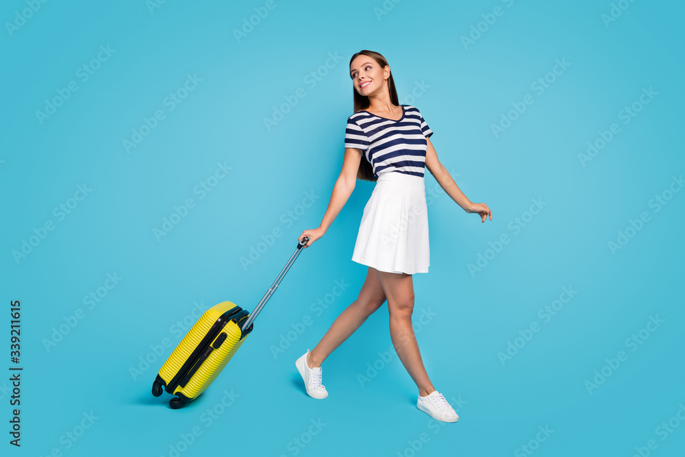 Full body profile photo of beautiful lady traveler walking flight registration rolling suitcase summer journey wear striped t-shirt white short skirt shoes isolated blue color background Full body profile photo of beautiful lady traveler walking flight registration rolling suitcase summer journey wear striped t-shirt white short skirt shoes isolated blue color background