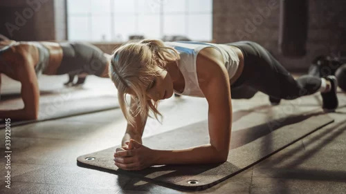Obraz Two Young Fit Atletic Women Hold a Plank Position in Order to Exercise Their Core Strength. They are Exhausted and Struggling with Training. They Workout in a Loft Gym.