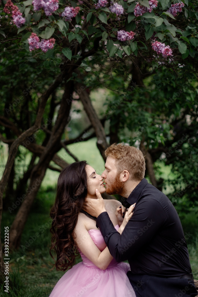 Beautiful couple in love cuddling in a blooming lilac garden. A woman in a purple sleeveless dress with a delicate necklace around her neck. Red-haired man in a black suit kisses and hugs a girl Beautiful couple in love cuddling in a blooming lilac garden. A woman in a purple sleeveless dress with a delicate necklace around her neck. Red-haired man in a black suit kisses and hugs a girl