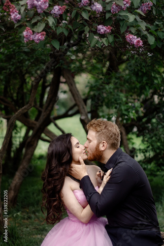 Beautiful couple in love cuddling in a blooming lilac garden. A woman in a purple sleeveless dress with a delicate necklace around her neck. Red-haired man in a black suit kisses and hugs a girl