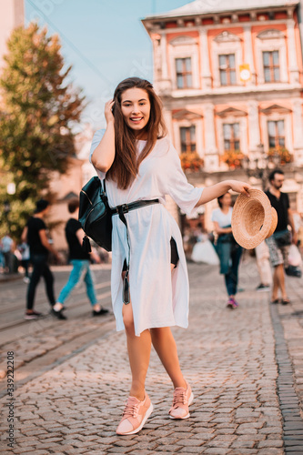 Young stylish woman walking on the old town street, travel with backpack, straw hat, wearing trendy outfit. © zadorozhna