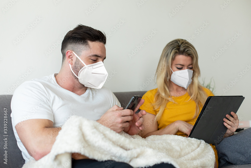 Couple wearing face mask using digital devices under Covid-19 quarantine. Digital leisure at home under coronavirus worldwide crisis,