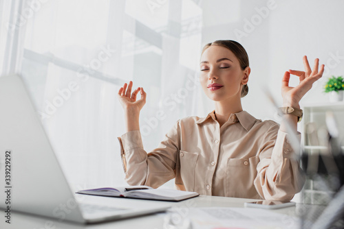 Beautiful businesswoman meditating with closed eyes and gyan mudra at workplace with laptop
