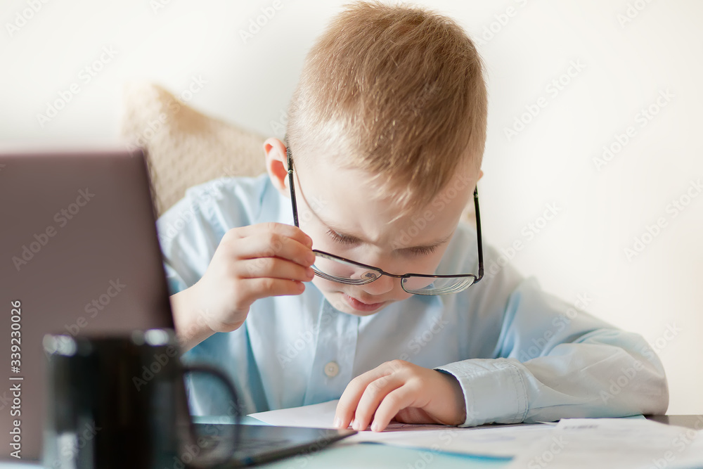 Workspace office desk with laptop and cup of coffee. Little cute business boy sitting at table and looking on a important documents. Kid learning online. Workspace office desk with laptop and cup of coffee. Little cute business boy sitting at table and looking on a important documents. Kid learning online.