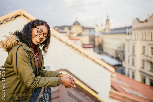 Positive playful black woman on the top of the building with european city view on background.