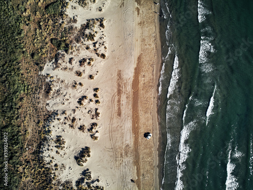 top aerial shot of a single car on the beach. Drone from above, blue water on sandy beach with white waves on the ocean.