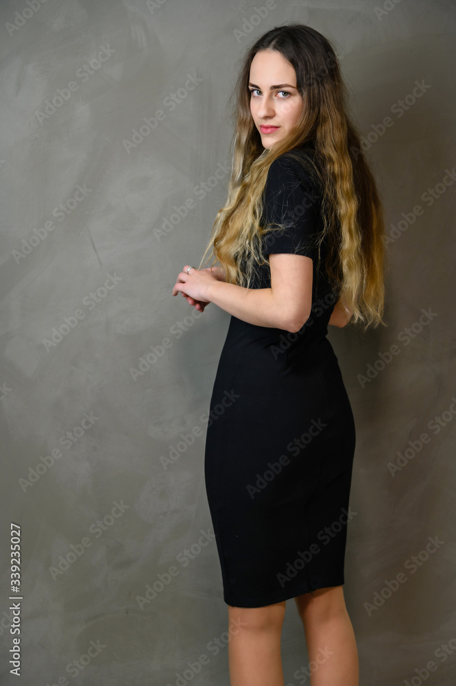 Vertical photo of a slender young Caucasian girl model with long curly hair in a black dress on a gray background. Made in the studio. Vertical photo of a slender young Caucasian girl model with long curly hair in a black dress on a gray background. Made in the studio.