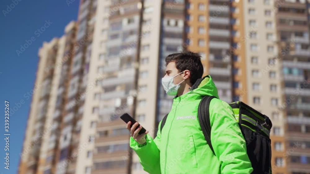Panning slow motion shot of young male food courier in medical mask using navigation app on cell phone while walking down street with insulated bag delivering order Panning slow motion shot of young male food courier in medical mask using navigation app on cell phone while walking down street with insulated bag delivering order