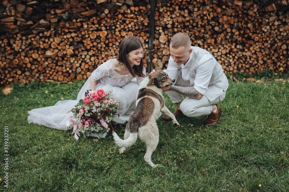 Spring wedding in the mountains. A guy in a shirt and vest and a girl in a white dress and with a bouquet in their hands are standing near a barn with firewood Spring wedding in the mountains. A guy in a shirt and vest and a girl in a white dress and with a bouquet in their hands are standing near a barn with firewood