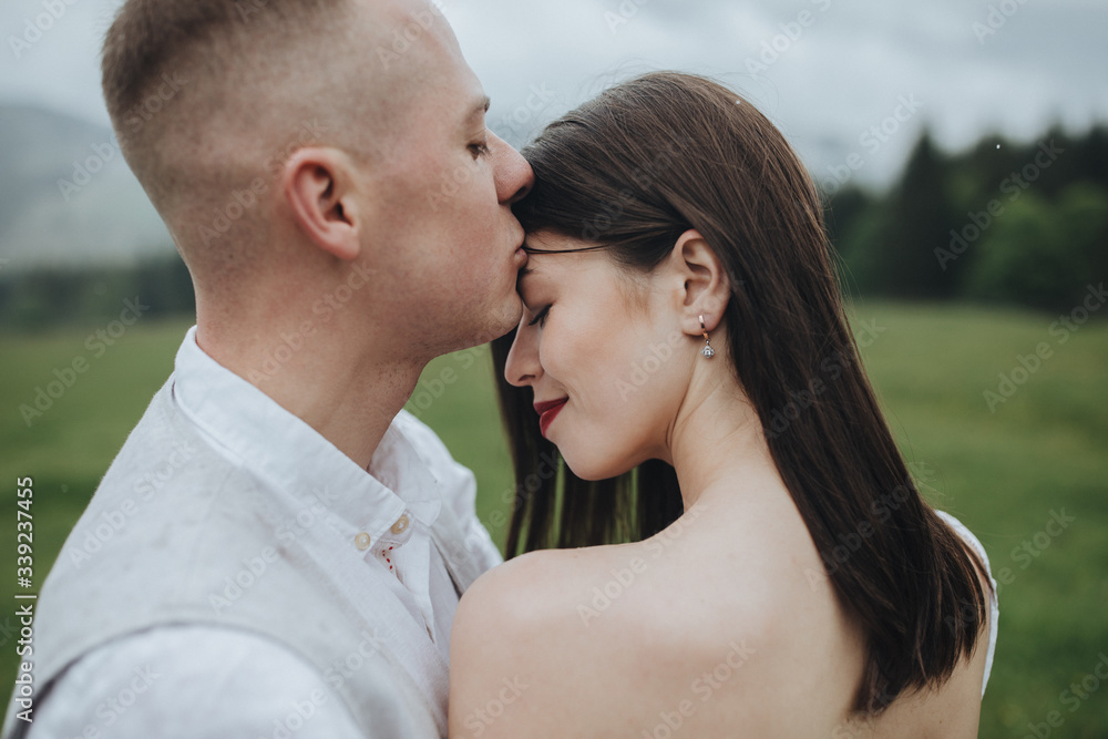 Spring wedding in the mountains. A guy in a shirt and vest and a girl in a white dress hug and kiss on a background of mountains and a forest Spring wedding in the mountains. A guy in a shirt and vest and a girl in a white dress hug and kiss on a background of mountains and a forest