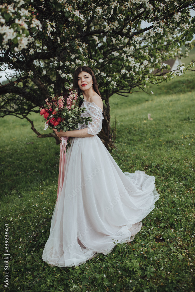 Spring wedding in the mountains. A young girl in a white dress stands near a blossoming tree in the mountains and holds in her hands a bouquet of red flowers with a ribbon Spring wedding in the mountains. A young girl in a white dress stands near a blossoming tree in the mountains and holds in her hands a bouquet of red flowers with a ribbon