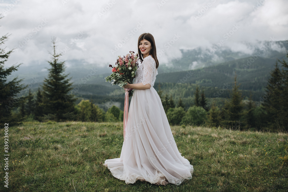 Spring wedding in the mountains. A young girl in a white dress stands in a green meadow in the mountains and holds in her hands a bouquet of red flowers and greenery