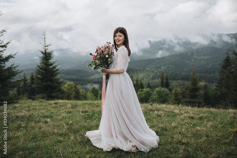 Spring wedding in the mountains. A young girl in a white dress stands in a green meadow in the mountains and holds in her hands a bouquet of red flowers and greenery Spring wedding in the mountains. A young girl in a white dress stands in a green meadow in the mountains and holds in her hands a bouquet of red flowers and greenery