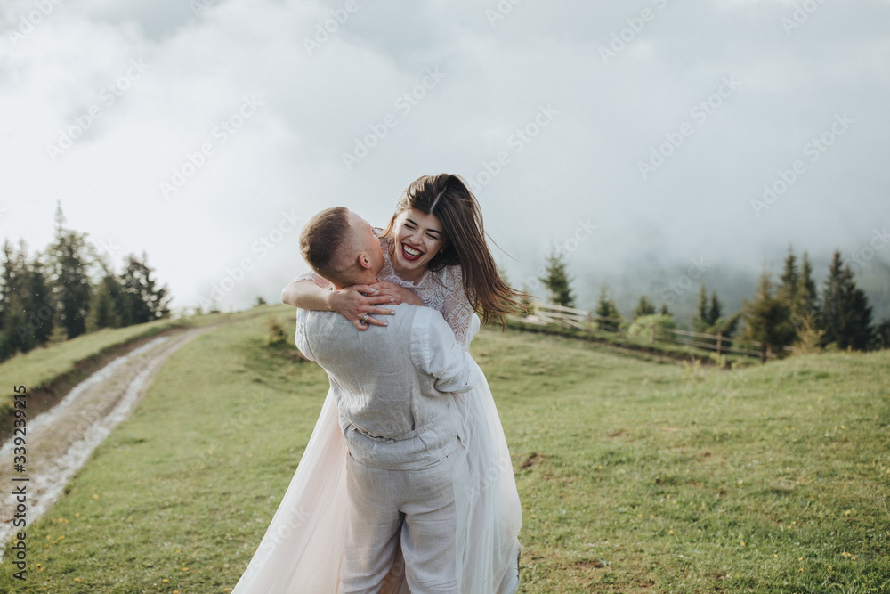 Spring wedding in the mountains. A guy in a shirt and vest and a girl in a white dress walk along a mountain road at sunset among the clouds