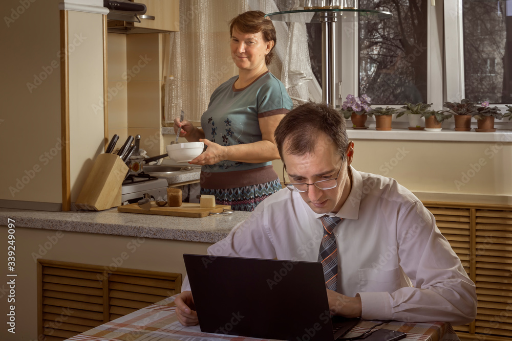 Young man works at a computer at home on the background of a woman busy cooking. Toned Young man works at a computer at home on the background of a woman busy cooking. Toned