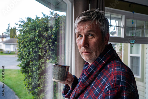 Unshaved middle aged man with messy hair holds coffee by window