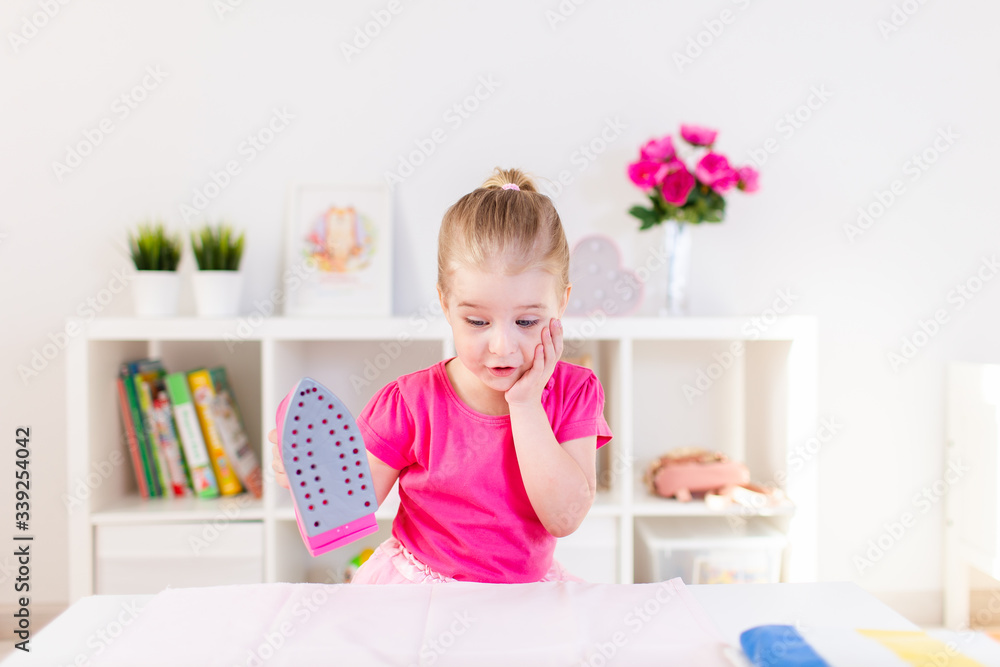 Little blonde baby girl housewife playing with toy pink iron. Ironong clothes on a white table. Housework, helper Little blonde baby girl housewife playing with toy pink iron. Ironong clothes on a white table. Housework, helper