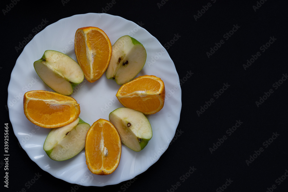 Slices of apple and orange on white plate on dark black background, top view Slices of apple and orange on white plate on dark black background, top view