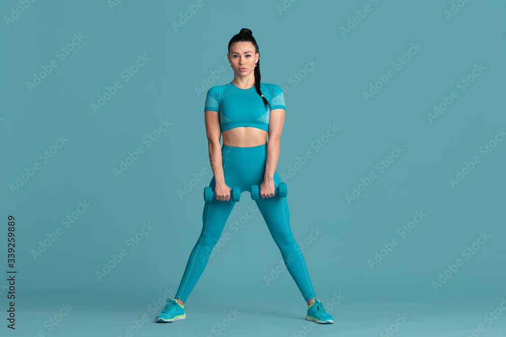 Graceful. Beautiful young female athlete practicing in studio, monochrome blue portrait. Sportive fit brunette model with weights. Body building, healthy lifestyle, beauty and action concept.