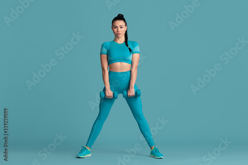 Graceful. Beautiful young female athlete practicing in studio, monochrome blue portrait. Sportive fit brunette model with weights. Body building, healthy lifestyle, beauty and action concept.