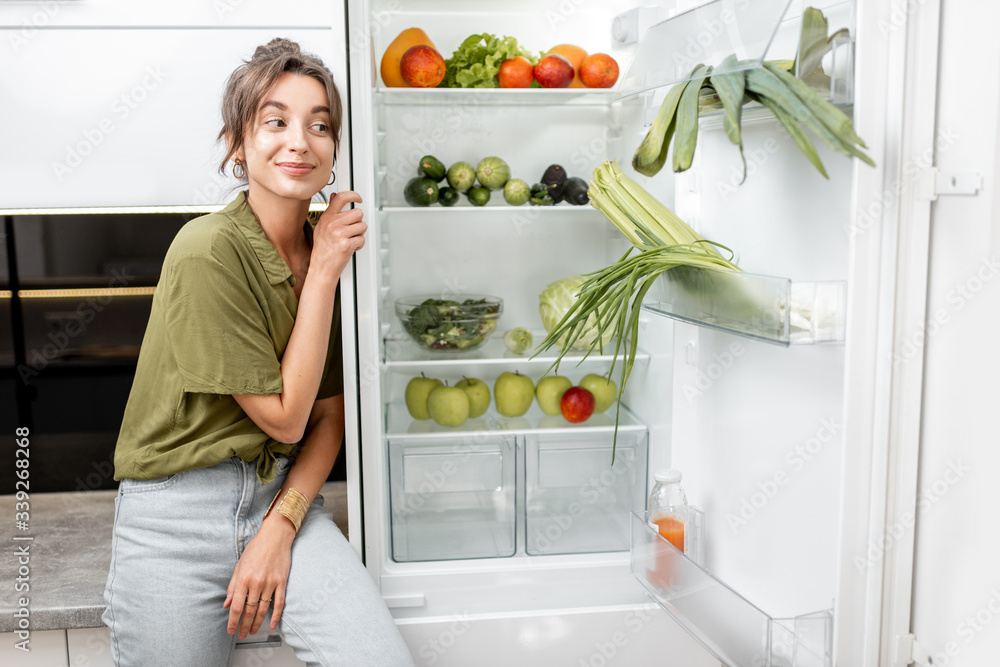 Portrait of a young woman sitting on the kitchen table near the fridge full of fresh and healthy food at home. Vegetarianism, wellbeing and healthy lifestyle concept Portrait of a young woman sitting on the kitchen table near the fridge full of fresh and healthy food at home. Vegetarianism, wellbeing and healthy lifestyle concept