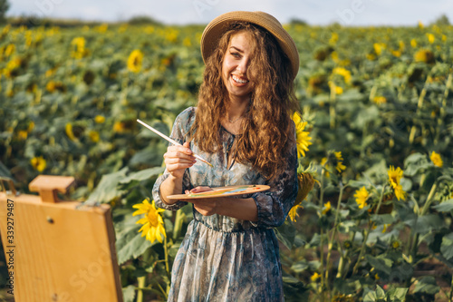A young woman with curly hair and wearing a hat is painting in nature. A woman stands in a sunflower field on a beautiful day