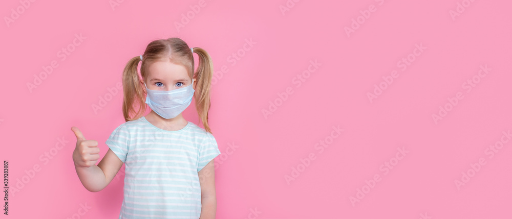 Little blonde girl with two ponytales in a medical mask face on a pink background showing thumbs up. Copyspace.