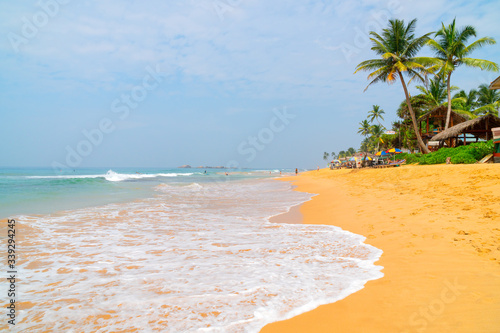 Hikkaduwa, Sri Lanka. March 8, 2018. Beach on the Indian Ocean. Sunny day, yellow sand, palm trees and foam waves.