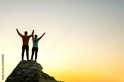 Man and woman hikers standing on a big stone at sunset in mountains. Couple raising up hands on high rock in evening nature. Tourism, traveling and healthy lifestyle concept.