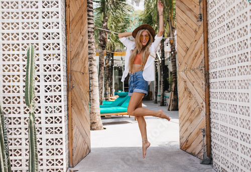 Full-length portrait of jumping blonde woman in yellow sunglasses laughing to camera. Outdoor photo of romantic girl expressing happiness chilling at summer resort.