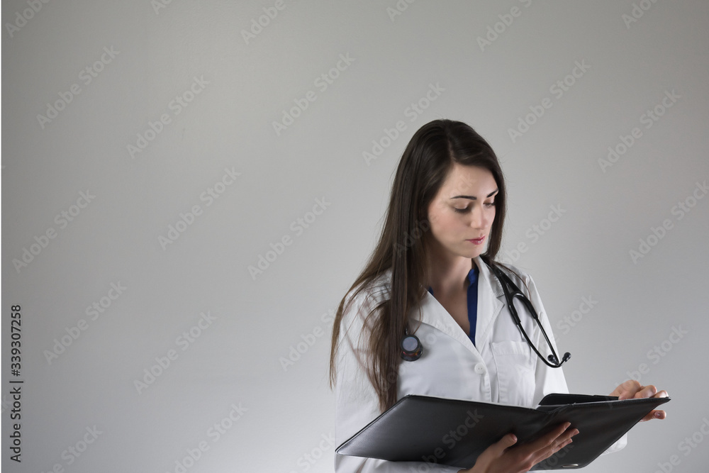 Female healthcare professional examining patient chart isolated on grey background. Stethoscope around neck, white coat on and buttoned. Female healthcare professional examining patient chart isolated on grey background. Stethoscope around neck, white coat on and buttoned.