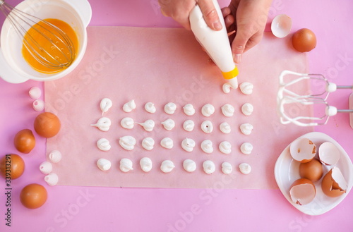 The process of making meringue cookies step by step. In the hands of a cooking bag for cream, squeezes out the meringue. On a pink background, parchment paper for baking, eggs, hand mixer, whisk.