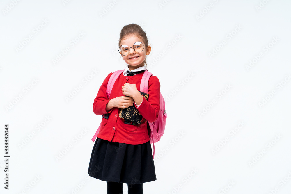 Adorable little girl in red school jacket, black dress, backpack and rounded glasses hugs a book tightly and smiles while enjoying and posing on white studio background. Isolate. Adorable little girl in red school jacket, black dress, backpack and rounded glasses hugs a book tightly and smiles while enjoying and posing on white studio background. Isolate.