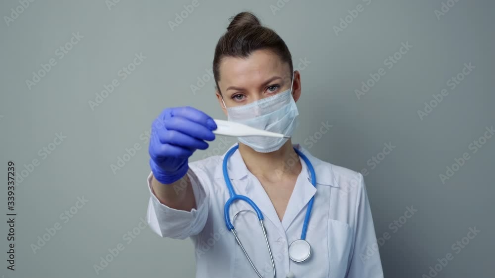 A female medical worker in a white robe and mask holds and looks at an electronic thermometer. Measuring temperature. A female medical worker in a white robe and mask holds and looks at an electronic thermometer. Measuring temperature.