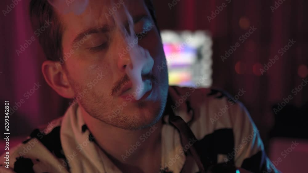 close-up of a man Smoking a pipe.a man with a mustache and stubble.the room has colored lighting, pink and blue.He is sitting on a stool at the table.seductively opinion and thoughts.a lot of smoke close-up of a man Smoking a pipe.a man with a mustache and stubble.the room has colored lighting, pink and blue.He is sitting on a stool at the table.seductively opinion and thoughts.a lot of smoke