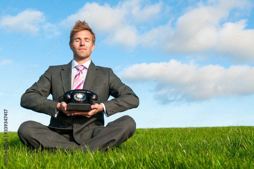 Meditating businessman sitting outdoors in sunny field holding an old-fashioned rotary dial phone Meditating businessman sitting outdoors in sunny field holding an old-fashioned rotary dial phone